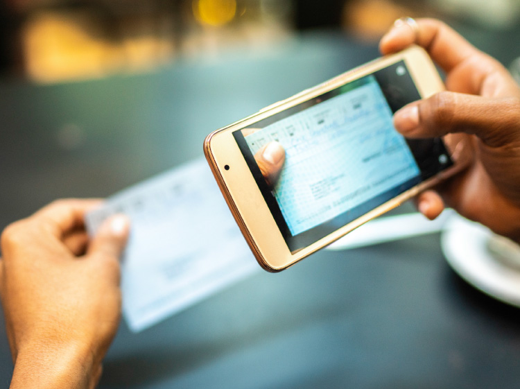 Close up of someone's hands holding a phone and a check. They are taking a picture of the check.