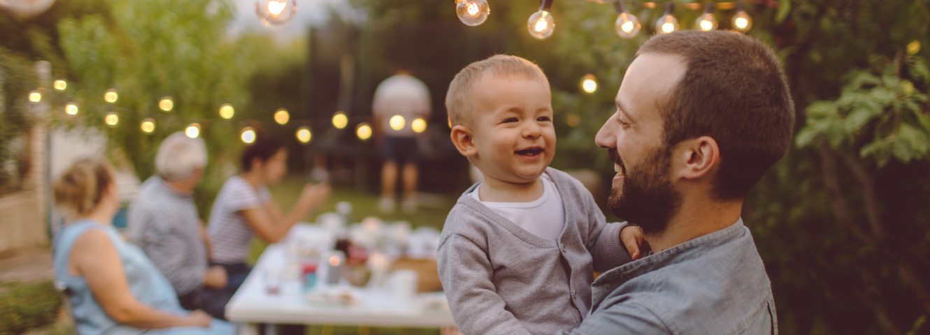 A dad holding his toddler. There are more family members behind them at a table.