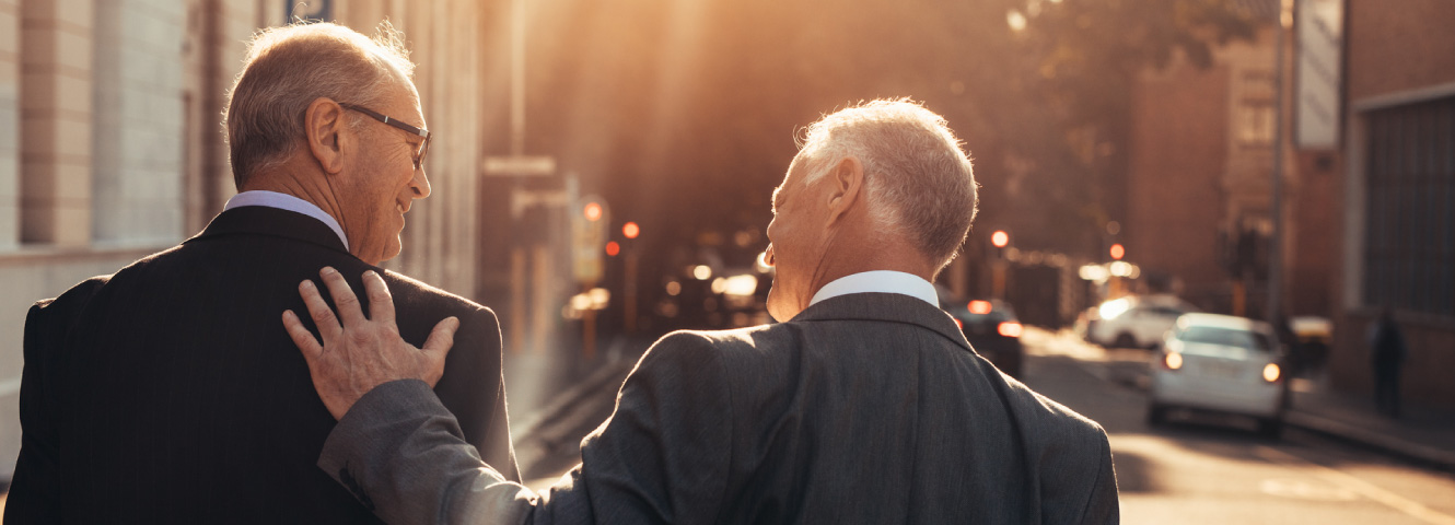 Two older businessmen in suits smiling at each other and walking down a city street.