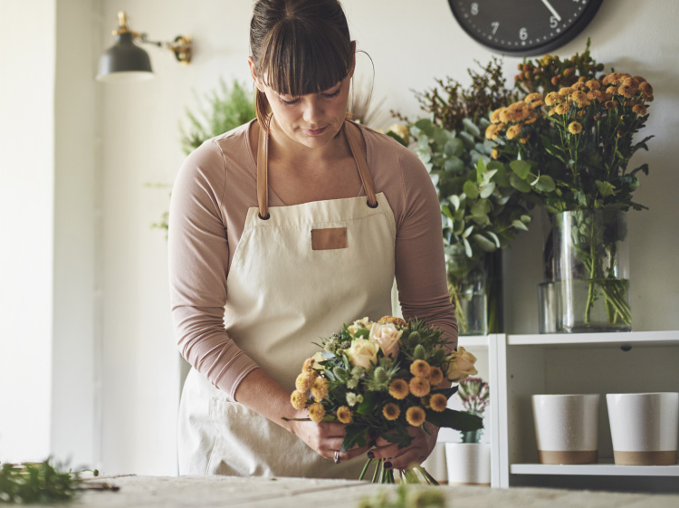 A young woman in an apron arranging flowers. There are more flowers in the background.