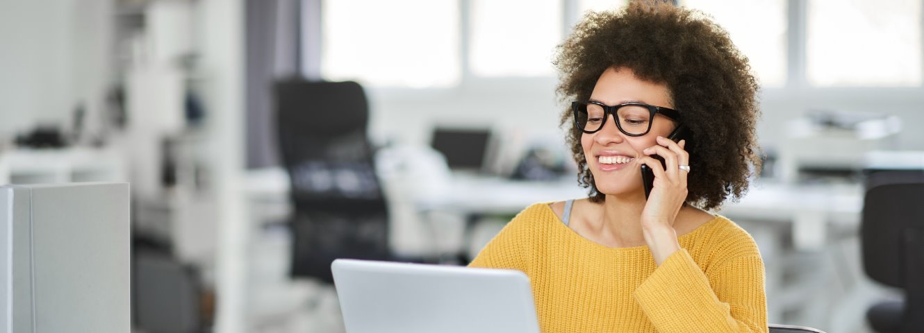 woman with glasses sitting at computer with phone in hand